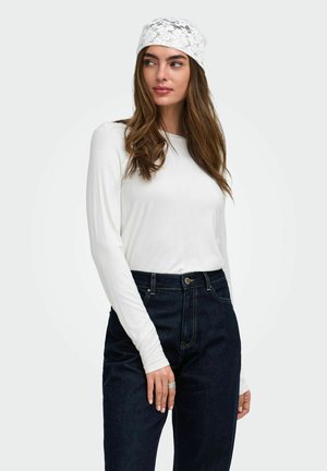 Femme aux longs cheveux bruns portant un foulard en dentelle blanche, un haut blanc à manches longues, un jean taille haute bleu foncé et une bague en argent à son doigt.