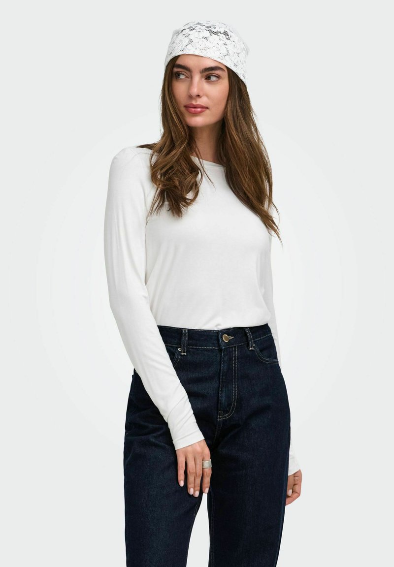 Femme aux longs cheveux bruns portant un foulard en dentelle blanche, un haut blanc à manches longues, un jean taille haute bleu foncé et une bague en argent à son doigt.