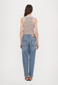 Vaqueros de mezclilla azul con un corte relajado, con una parche de cuero marrón en la cintura trasera y dos bolsillos traseros. Lleva puesta una camiseta de tirantes a rayas.