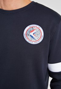 Sweat-shirt bleu marine avec un col rond côtelé, arborant un patch circulaire Apollo 15 avec des motifs rouges, bleus et gris, ainsi qu'une bande blanche sur la manche.