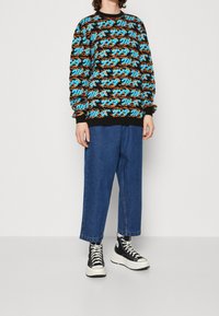Osoba ubrana w sweter w czarno-pomarańczowo-niebieski wzór, luźne niebieskie dżinsy skrócone do kostek oraz czarne wysokie sneakersy z białymi podeszwami.