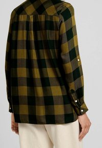 Chemise à manches longues à carreaux alternant des carrés verts et olive, avec des poignets boutonnés et un détail plissé dans le dos.