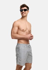 Grå svømmebukser med et bølgemønster, elastisk talje og hvid snor. Båret med sorte solbriller. Ingen skjorte eller yderligere accessories.