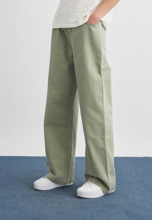 Henkilö, joka käyttää vaaleanvihreitä leveälahkeisia housuja ja valkoisia lenkkareita, seisoo sinisellä matolla käsi taskussa.