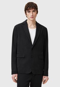 Schwarzer Blazer mit maßgeschneidertem Schnitt, zwei vorderen Taschen, Reverskragen und Zweiknopfverschluss. Getragen über einem weißen gerippten Hemd.