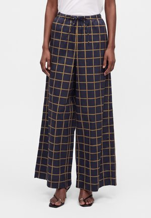 Pantalon large bleu marine à motif quadrillé marron et taille élastique avec cordon, porté avec des talons ouverts noirs sur une personne à la peau foncée.