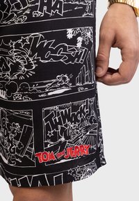 Svart shorts med et hvitt tegneserieprint av Tom og Jerry, med ulike lydeffekter i tegneseriestil; rødt brodert logo på kanten.