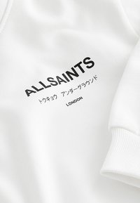 Fehér pulóver, élénk fekete szöveggel, rajta a "ALLSAINTS" és "LONDON" márkanevével, valamint japán karakterekkel. Rózsás mandzsetták.