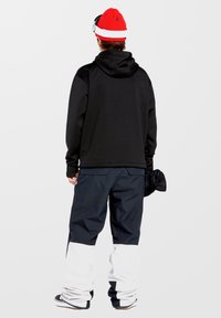 Schwarzer Kapuzensweater, dunkelblaue Hose mit weißen unteren Bündchen, rotes Beanie mit weißem Band und hält einen schwarzen Gegenstand in der Hand.