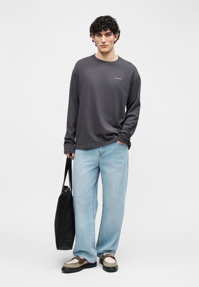 Νεαρός άνδρας που φοράει σκούρο γκρι μακρυμάνικο πουκάμισο, ανοιχτόχρωμα μπλε τζιν, δίχρωμα loafers, κρατώντας μια μαύρη τσάντα tote με το ένα χέρι στην τσέπη.