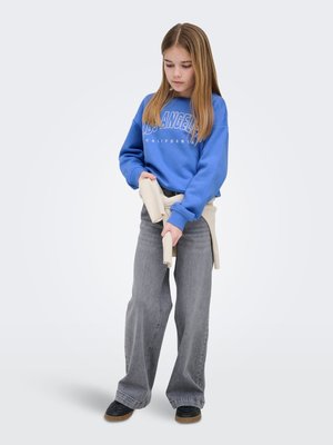 Fille aux cheveux lisses châtain clair portant un sweat-shirt bleu Los Angeles, un jean gris large, des chaussures noires et tenant un pull beige autour de la taille.