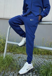 Mørkeblå træningsdragt med en lynlåset jakke, der har et logo og sidelommer, parret med hvide Nike-sko med sorte såler.