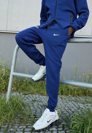 Navy blue tracksuit with a zippered jacket, featuring a logo and side pockets, paired with white Nike shoes with black soles.