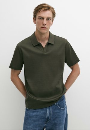 Junger Mann mit hellbraunen Haaren, der ein olivgrünes kurzärmeliges Polohemd und blaue Jeans trägt, vor weißem Hintergrund stehend.
