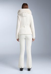 Fehér dzseki műszőrme kapucnival, testhezálló kialakítással, övvel a deréknál, és hosszú ujjakkal. Hozzáillő trapéznadrággal, cipzáras részletekkel.