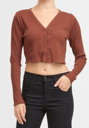 Femme portant un haut court côtelé couleur rouille à manches longues boutonné et un jean noir taille haute, main dans la poche.