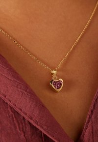 Gouden hart hanger ketting met roze edelsteen accenten, bevestigd aan een fijne gouden ketting, gedragen tegen een huidkleurige achtergrond.