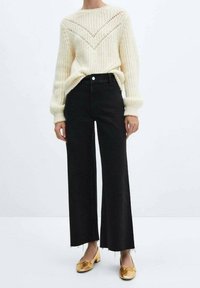 Suéter blanco de punto con un patrón texturizado, jeans de pierna ancha negros de altura alta y bailarinas doradas, con detalles de dobladillo deshilachado.