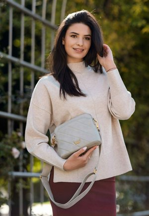Graue gesteppte Handtasche mit goldenem Logo und langem Tragegurt, positioniert gegen einen hellbeigen Pullover und einen burgunderfarbenen Rock, im Freien mit Grünpflanzen im Hintergrund.