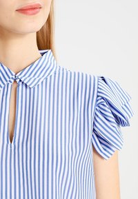 Modrá a bílá pruhovaná blůza s límečkem, detaily v oblasti klíčov é dírky a volnými rukávy. Vyrobena z lehké tkaniny s hladkou texturou.
