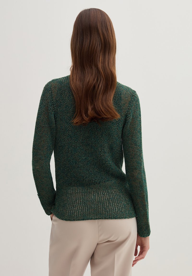 Suéter de punto en verde oscuro con un patrón texturizado, mangas largas y un dorso ligeramente alargado. Combinado con pantalones beige claros.