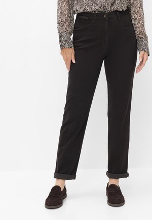 Vrouw draagt zwarte, rechte opgerolde broekspijpen, een patroon lange mouwen shirt en donkerbruine slip-on loafers, staand tegen een witte achtergrond.