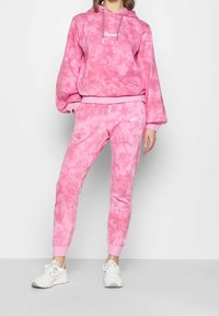 Kobieta w różowej bluzie tie-dye i dopasowanych dresach w tym samym kolorze, w białych trampkach, stojąca na tle jasnej, jednolitej ściany.