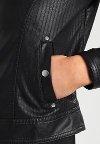 Sort læderjakke med ribbet tekstur, en sidelomme og sølvfarvede trykknapdetaljer på manchetten. Glat overflade med diskret glans.