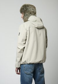 Veste beige à capuche avec une texture lisse et un chapeau doublé en fourrure. Présente des poignets élastiques et un logo sur la manche gauche. Portée avec un jean bleu.