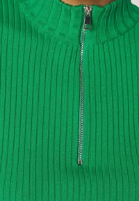 Gros plan sur un pull vert à côtes avec une fermeture éclair argentée partiellement ouverte partant du col montant vers la poitrine.