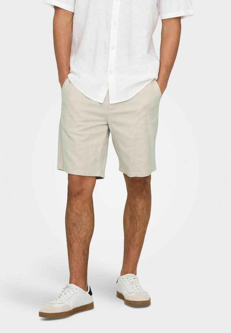 Light beige shorts made of a soft fabric, featuring side pockets and a straight cut, paired with white sneakers and a white short-sleeve shirt.
