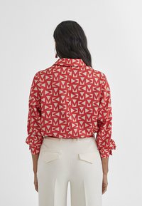 Blusa rossa a fantasia con forme geometriche in crema. Maniche lunghe con polsini arrotolati, colletto con bottoni e design dritto sul retro.