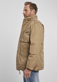 Jachetă khaki cu guler înalt, mai multe buzunare frontale și închidere cu fermoar, având o textură moale a materialului și o croială relaxată.