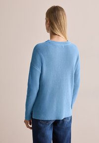 Blå stickad tröja med ribbat mönster, rund halsringning och sidofickor. Baksidan har en avslappnad passform och sänkta axelsömmar.