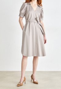 Beige mekko, jossa on V-pääntie, puffihihat, vyötärönauha ja polvimittainen helma. Yhdistetty ruskeisiin kärkitossuihin neutraalilla taustalla.