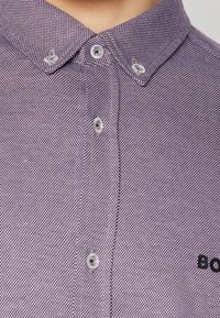 Chemise à manches courtes avec boutons, à motif à carreaux violet et blanc. Elle comporte un col, cinq boutons et un texte brodé sur la poitrine.
