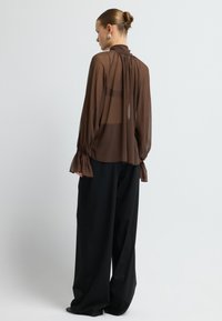 Frau mit einem Haarknoten, die eine transparente braune Bluse mit Glockenärmeln und lockeren schwarzen weiten Hosen trägt, steht vor einem schlichten hellen Hintergrund.