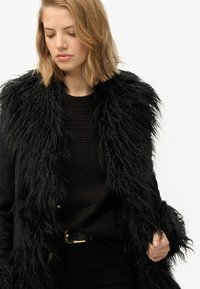 Sort faux fur frakke med en tyk, tekstureret krave. Båret over en mørk sweater med et glat stof og en bæltestrop i taljen. Minimalistisk design.