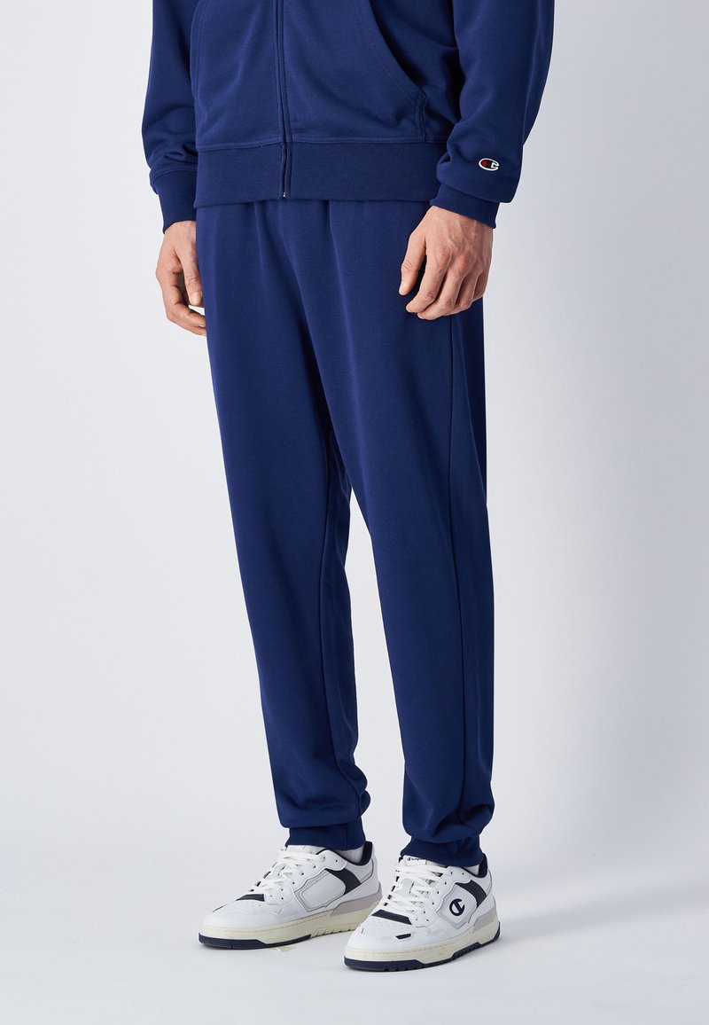 Marinefarbener Trainingsanzug mit einer Jacke mit Reißverschluss und passenden Hosen. Hergestellt aus weichem Stoff, mit elastischen Bündchen und Seiten. Weiße Sneaker mit schwarzen Akzenten.