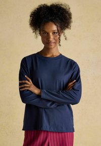 Marineblaues Langarmshirt aus weichem Stoff, mit Rundhalsausschnitt und lockerer Passform. Das Model posiert mit verschränkten Armen, der Hintergrund ist leicht strukturiert.