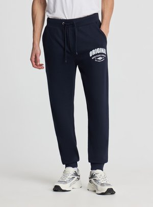 Mężczyzna w granatowych spodniach dresowych z białym napisem „ORIGINAL Heritage Denim” na lewym udzie i białych trampkach.