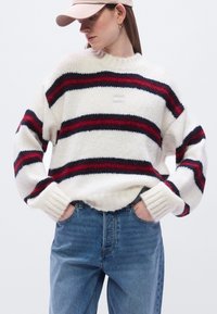 Χοντρή πλεκτή πουλόβερ σε κρεμ απόχρωση με κόκκινες και ναυτικές οριζόντιες ρίγες, στρογγυλή λαιμόκοψη και ριμπ περιλαίμια, συνδυασμένο με ανοιχτόχρωμα τζιν.
