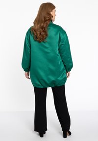 Groene satijnen oversized jas met elastische zoom en manchetten, gecombineerd met zwarte wijde broek en hakken, gezien vanuit de achterkant.