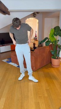 Młody mężczyzna poprawiający szare spodnie dresowe w salonie z pomarańczową kanapą, dużą zieloną rośliną i drewnianą podłogą.