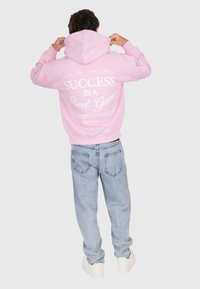 Persona che indossa un cappuccio rosa con la scritta "Il successo è un gioco di strada" e jeans azzurri chiari, con il cappuccio alzato, in piedi contro uno sfondo semplice.