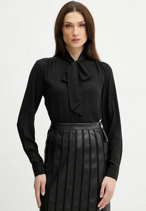 Donna che indossa una blusa nera a maniche lunghe con cravattino e una gonna nera a pieghe in pelle a vita alta, in piedi davanti a uno sfondo semplice.