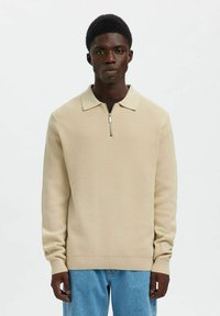 Beżowy sweter o fakturowanej powierzchni z kołnierzem i półzamkiem, z długimi rękawami i prążkowanymi mankietami. Połączony z jasnoniebieskimi dżinsami.