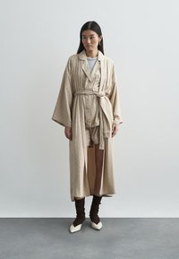 Μπεζ ρόμπα με σχέδιο, συνοδευόμενη από πουκάμισο και σορτς στο ίδιο χρώμα, με φαρδιά μανίκια και ζώνη στη μέση. Το μοντέλο φορά καφέ κάλτσες μέχρι το γόνατο και λευκά παπούτσια.