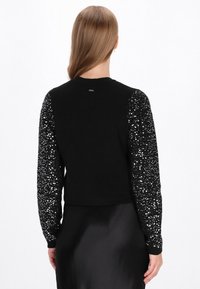 Fekete pulóver, flitterekkel díszített ujjal. Testhezálló fazon, kötött anyag, puha textúra, rövid hossza, bordás mandzsetták.