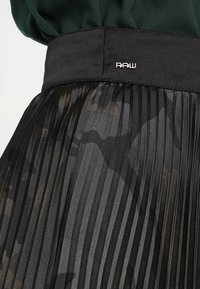 Gonna nera plissettata con una finitura lucida e semitrasparente, caratterizzata da un motivo camouflage. Ampio cinturino nero con dettagli del logo "RAW" in argento.
